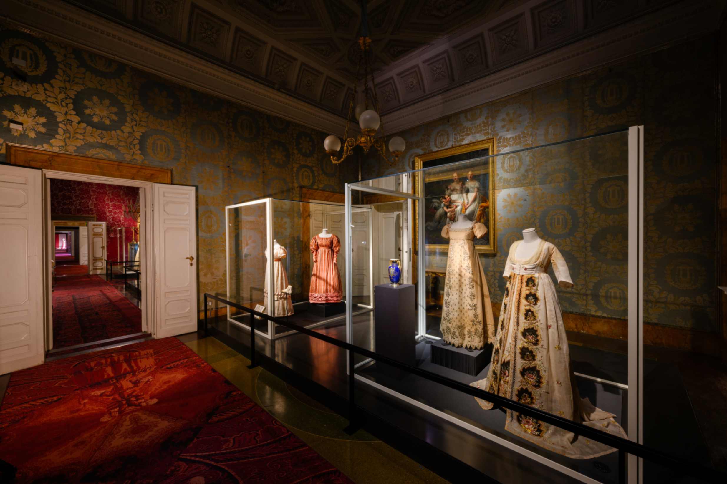 SALA 2: DAL NEOCLASSICISMO ALLA RESTAURAZIONE Veduta d’insieme di quattro abiti dal 1790 al 1825 ca. di manifattura italiana, un prezioso vaso (Manifattura di Vienna 1790) e alcuni accessori coevi. Sulla parete il dipinto Madame Rouillard con una sorella e un’amica di Jean-Sébastien Rouillard (Primo quarto del XIX secolo)