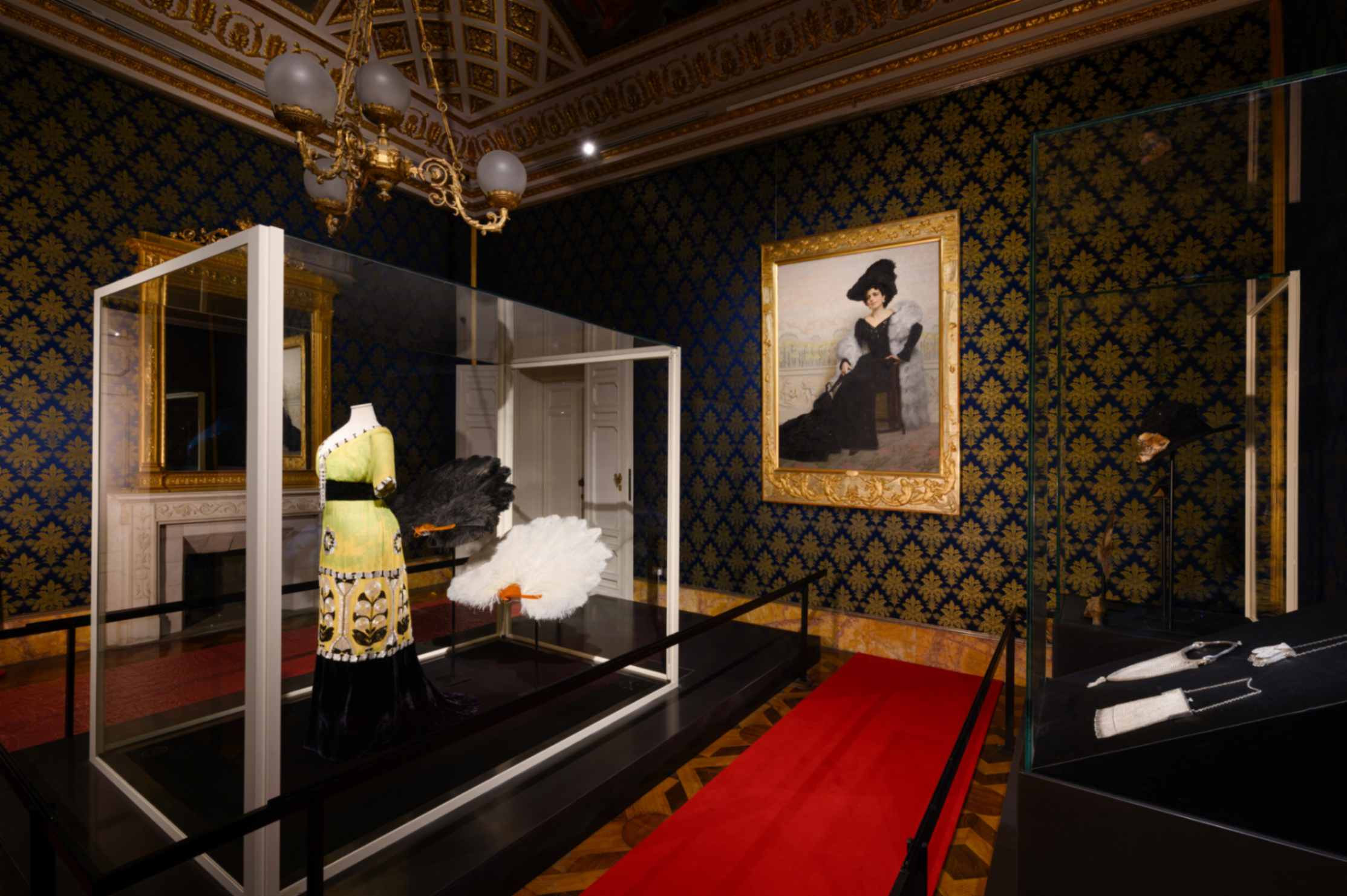 SALA 6 I PRIMI ANNI DEL NOVECENTO Veduta d’insieme con il prezioso abito liberty in chiffon di seta nelle tonalità del verde e del giallo con decorazioni-gioiello di Raphael Goudstikker (1913), due grandi ventagli in piume di struzzo, alcuni accessori coevi e, sulla parete, il Ritratto della signora Angiola Maria Pagliano-Bruno di Edoardo Gelli (1904)